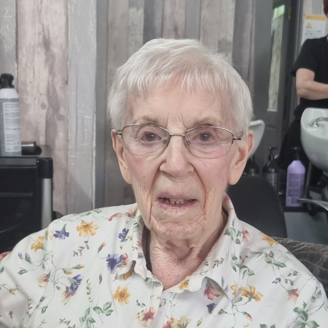 An elderly woman with freshly cut hair, wearing glasses and a floral blouse, sitting in a hair salon. The background features salon equipment and another person working.