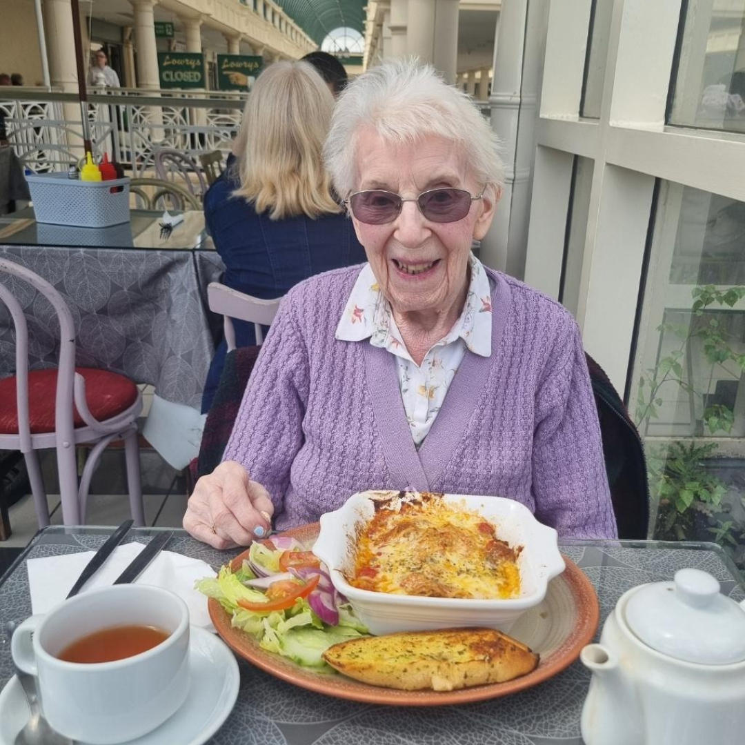 An elderly woman wearing a lavender cardigan and glasses, smiling while seated at a table in a café. She has a cup of tea and a plate of food including lasagne, salad, and garlic bread. The background features a busy café scene.