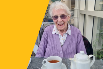 An elderly woman wearing a lavender cardigan and glasses, smiling while sitting at a table set with a teapot, teacup, and saucer. The website header graphic has a yellow frame around the image.