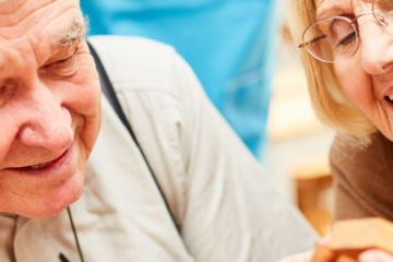 elderly man wearing a grey shirt with grey hair and an elderly woman wearing a brown jumper with blonde hair, sat looking down at something together, whilst smiling.